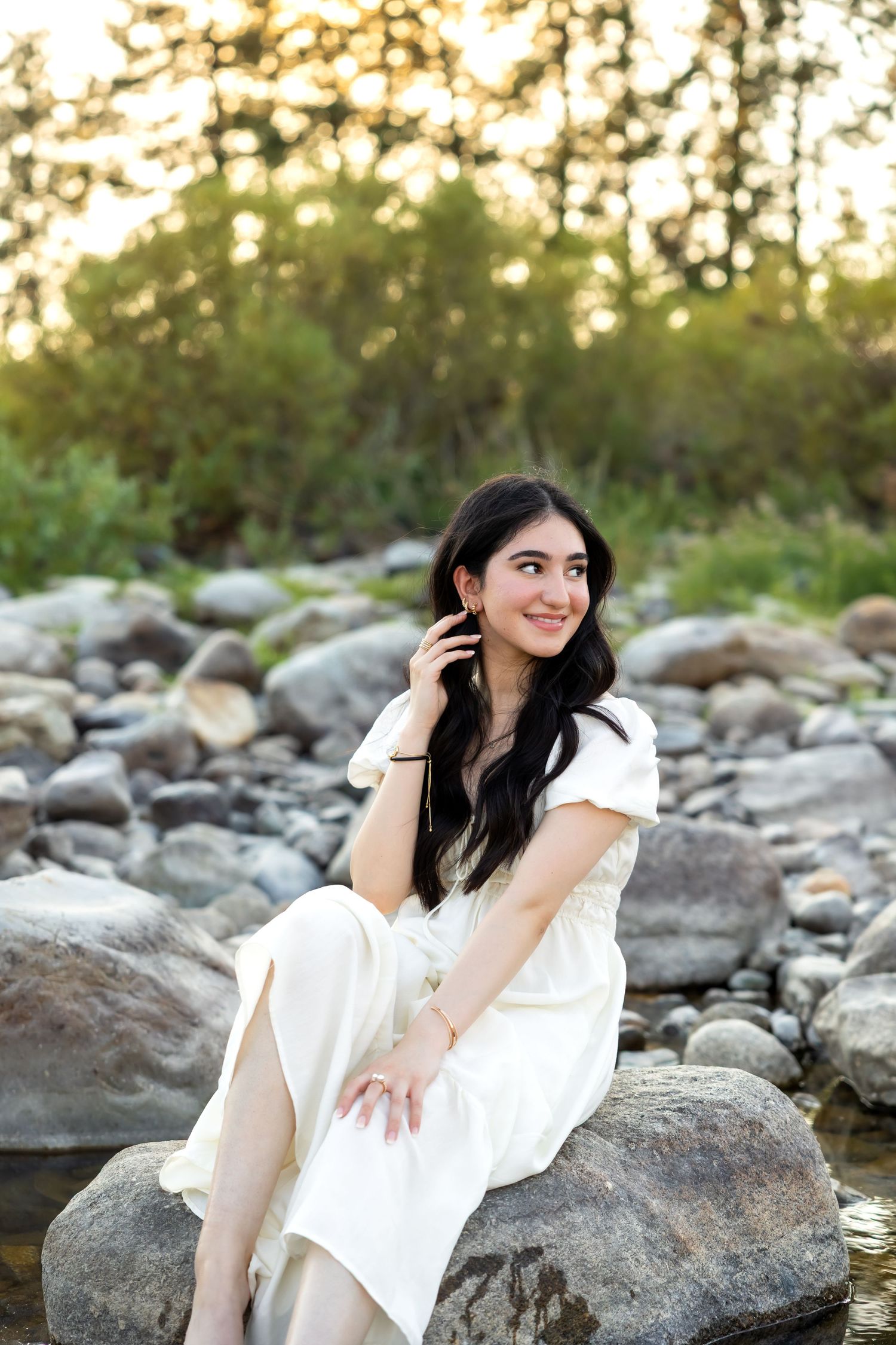 A person in a white dress poses gracefully on rocky terrain at sunset with trees in the background.