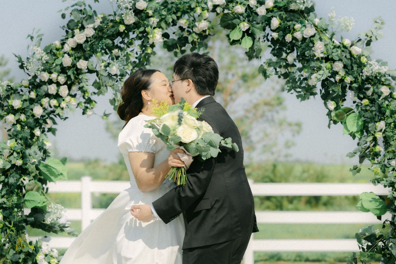 bride and groom kissing beneath a floral arch during an outdoor wedding ceremony