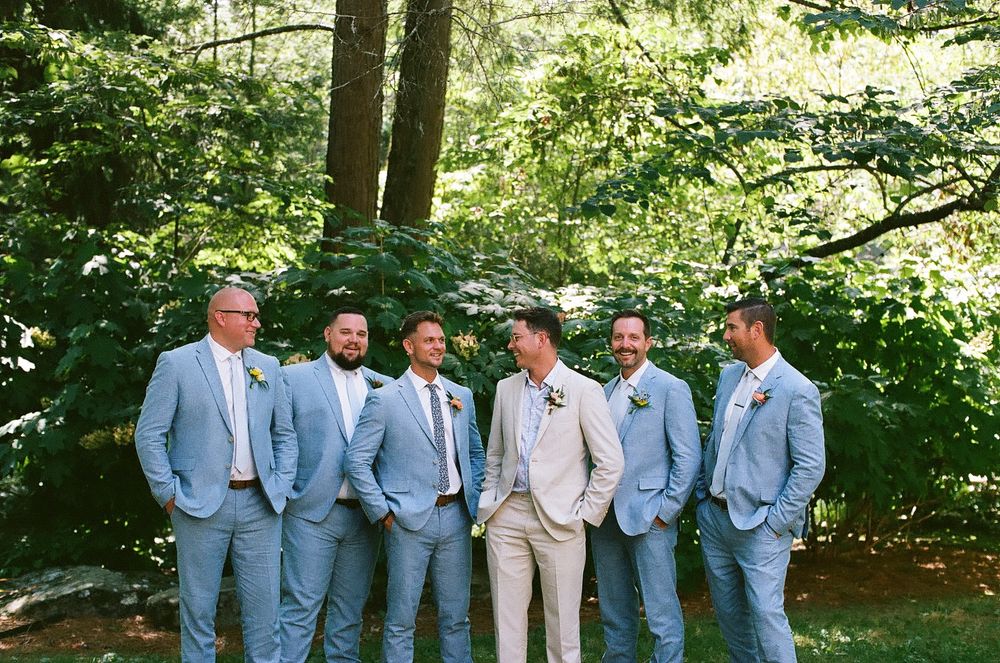 A group of groomsmen in light blue suits pose together in a wooded outdoor setting for wedding photos.