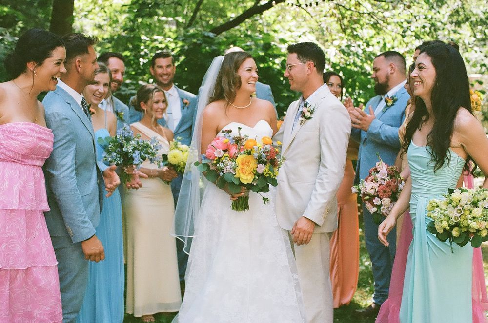 Wedding party in pastel blue and pink dresses poses together outdoors in a garden setting with lush greenery.