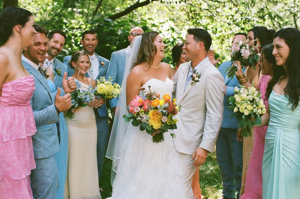 Wedding party in pastel dresses and light blue suits celebrates outdoors with colorful spring bouquets.