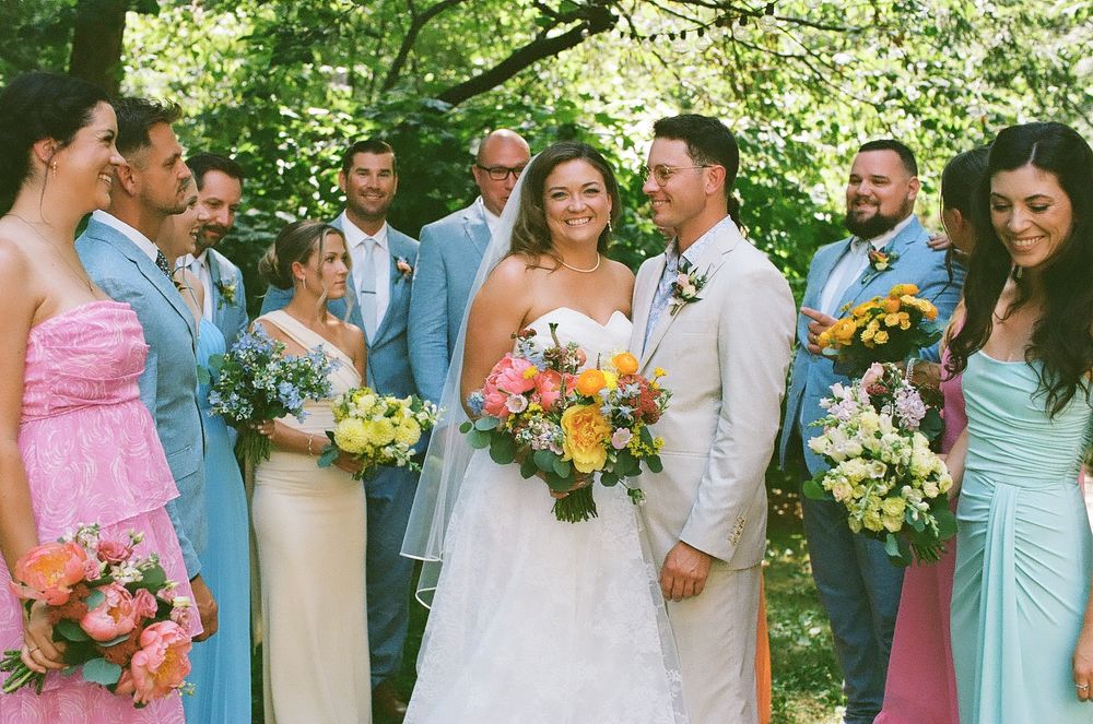 A wedding party in pastel blue and pink attire poses together outdoors surrounded by lush greenery and bright flowers.