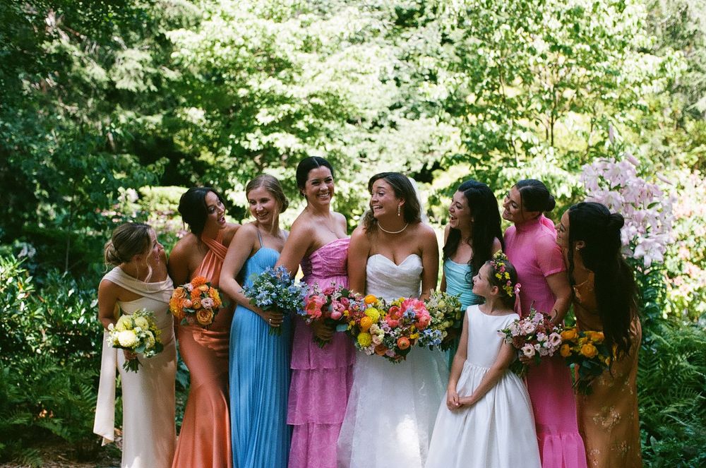A group in colorful dresses stands together holding vibrant floral bouquets in an outdoor garden setting.