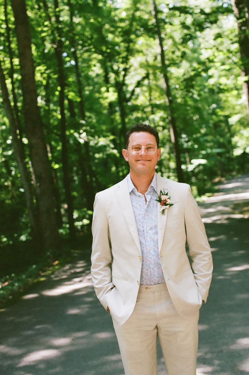 A person in a cream-colored suit stands on a wooded path surrounded by lush green trees on a sunny day.