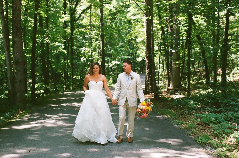 A bride and groom stroll hand in hand down a tree-lined path dappled with summer sunlight.