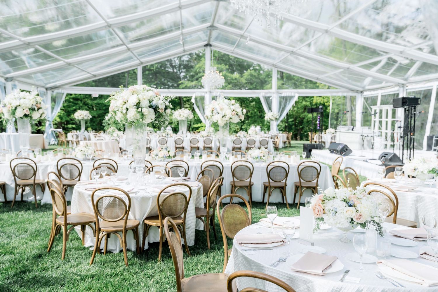 A luxurious outdoor wedding reception setup under a clear tent with elegant white decor and round tables.