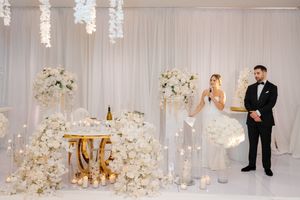 An elegant white wedding reception setup with tall floral arrangements and gold accents against a white backdrop.