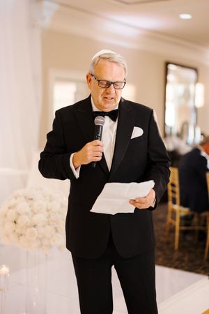 A speaker in a black suit and glasses delivers a speech while holding papers at an elegant indoor event.
