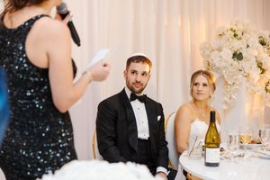 Wedding guests at a formal reception listen attentively to a toast speech while seated at an elegant table setting.