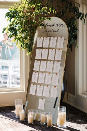 White seating chart display with greenery decor and glass candle holders arranged on a window ledge.