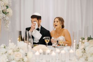 Wedding party sits at an elegant white reception table decorated with floral centerpieces and glowing candles.