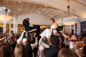 Wedding guests celebrate on a dance floor during a reception with arms raised under elegant chandeliers.
