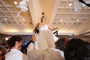 A wedding reception dance floor scene with guests celebrating under white ceiling tiles and warm lighting.