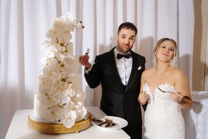 A wedding couple cuts into their elegant white and gold tiered cake decorated with cascading orchids at their reception.