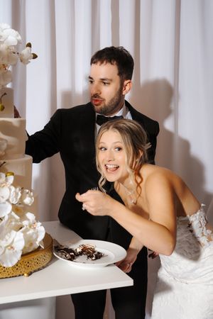 A couple in formal wedding attire playfully interacts at a white table decorated with orchids in a studio setting.