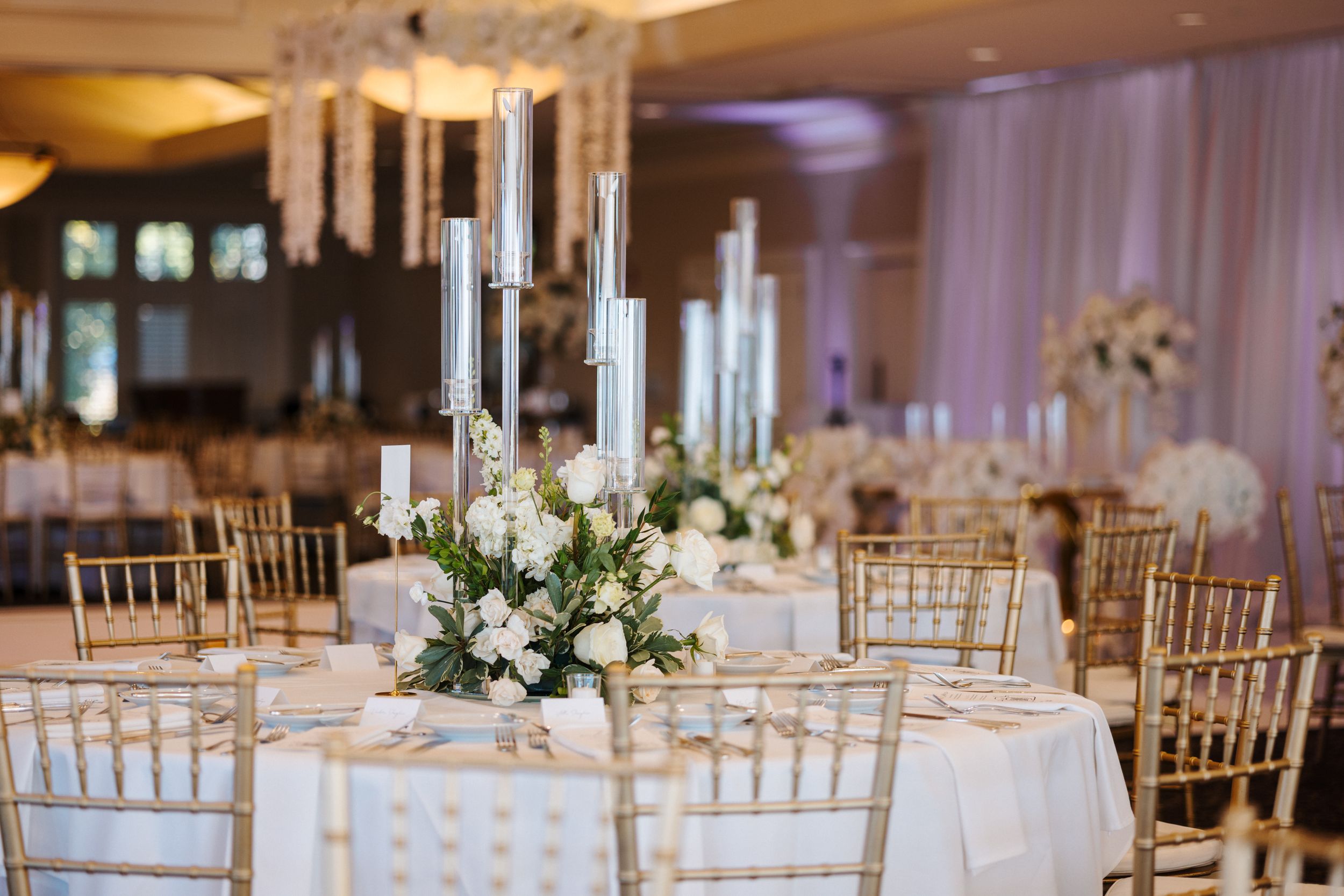 Elegant wedding reception table with white floral centerpiece, gold chiavari chairs and tall white candles in crystal holders.