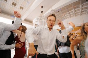 A group of people dancing and celebrating at a wedding reception in a white-walled banquet hall.