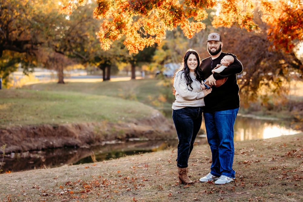 A couple poses together with their newborn in a park during autumn with golden foliage and warm sunlight in the background in Keller, TX