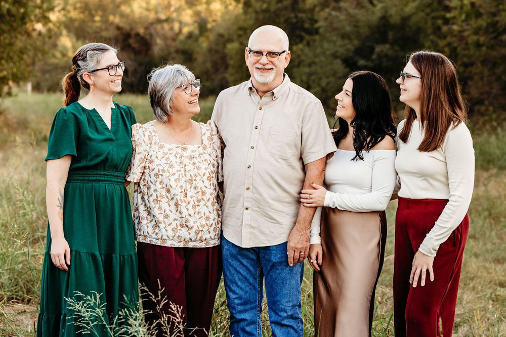 Parents with their three grown daughters in coordinated fall outfits stand together outdoors for a family portrait session in Fort Worth