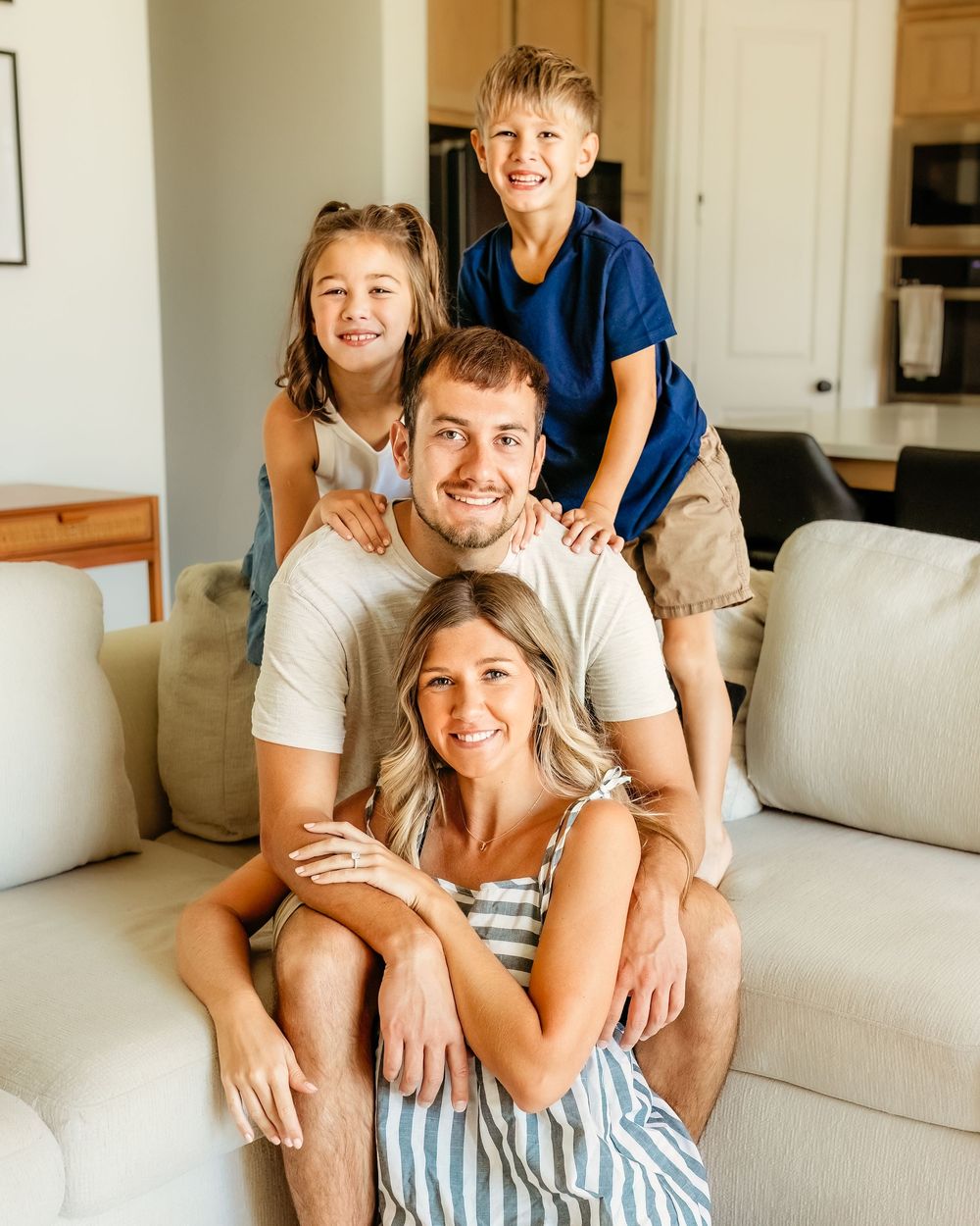 A family sits together on a light colored couch in a bright living room, sharing smiles and laughter during in-home session in DFW