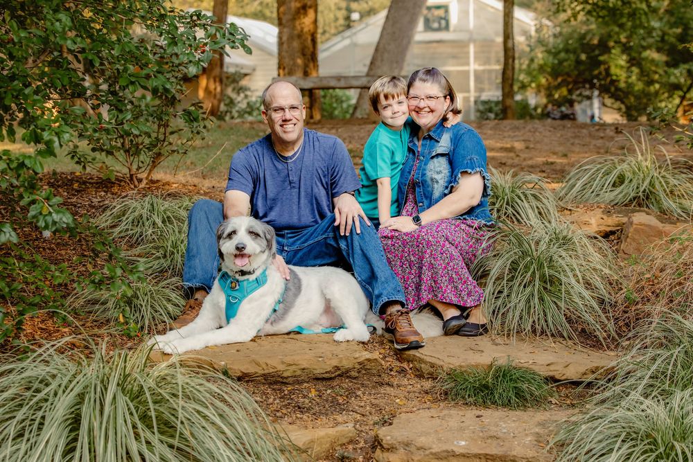 A family of three sits together on stone steps with their white dog in a landscaped garden setting during autumn in Grapevine, TX