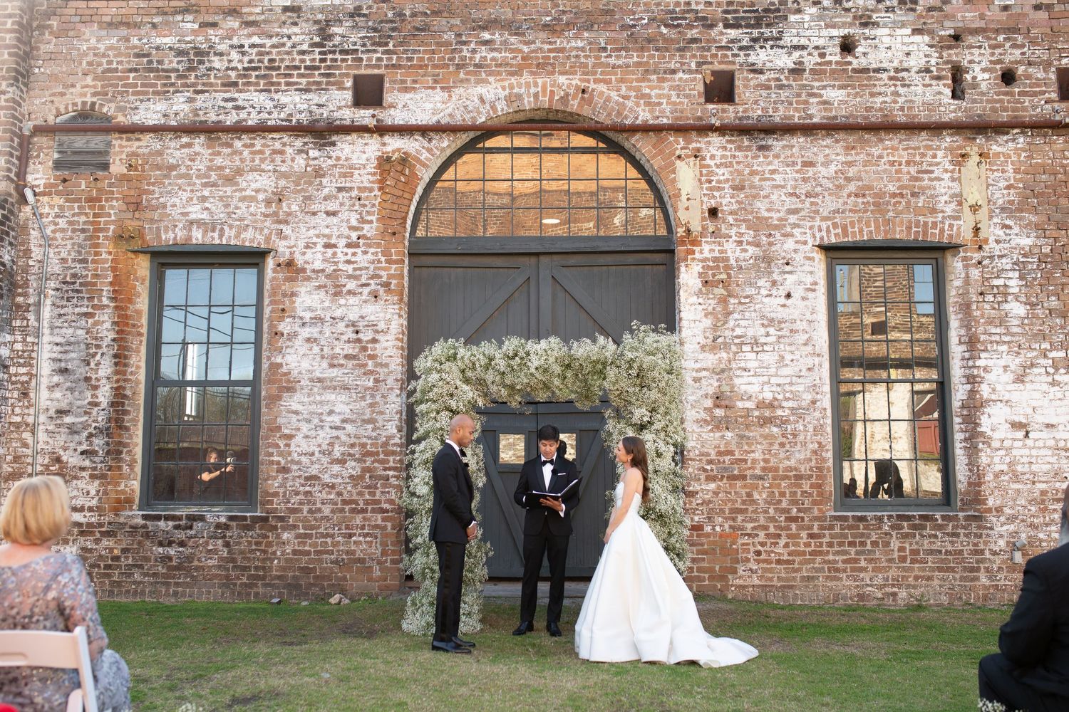 Indoor wedding ceremony with candle-lit aisle lined with greenery in a rustic industrial venue with exposed beams.