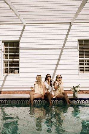 Three friends relaxing by a pool with their feet in the water against a white house exterior.
