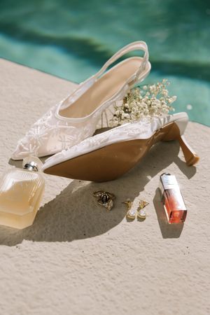 Wedding accessories including white lace heels, jewelry, and perfume arranged by a pool.