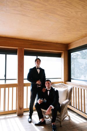 Two men in formal tuxedos pose on a wooden porch with large windows.