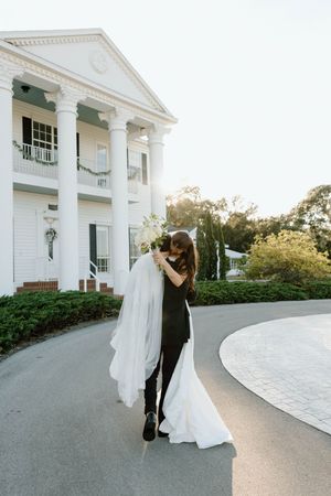 Newlyweds dance playfully along a curved driveway in front of a stately white mansion.