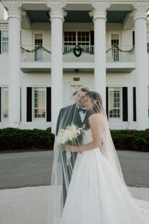 Elegant wedding couple embraces in front of a grand white colonial mansion with tall columns.