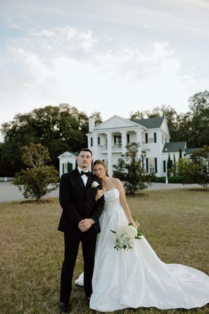 A wedding couple poses in front of a stately white colonial mansion with pillars and green lawn on their special day.