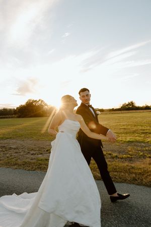 A couple in wedding attire dances playfully in a field at sunset while the bride's dress flows in the wind.
