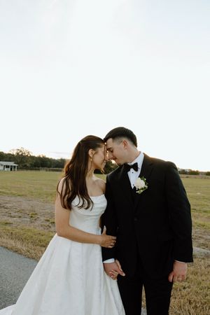A romantic outdoor wedding portrait of a couple in formal attire sharing an intimate moment at sunset.