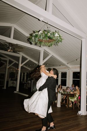Wedding couple performs dramatic dance dip on wooden floor under hanging greenery.