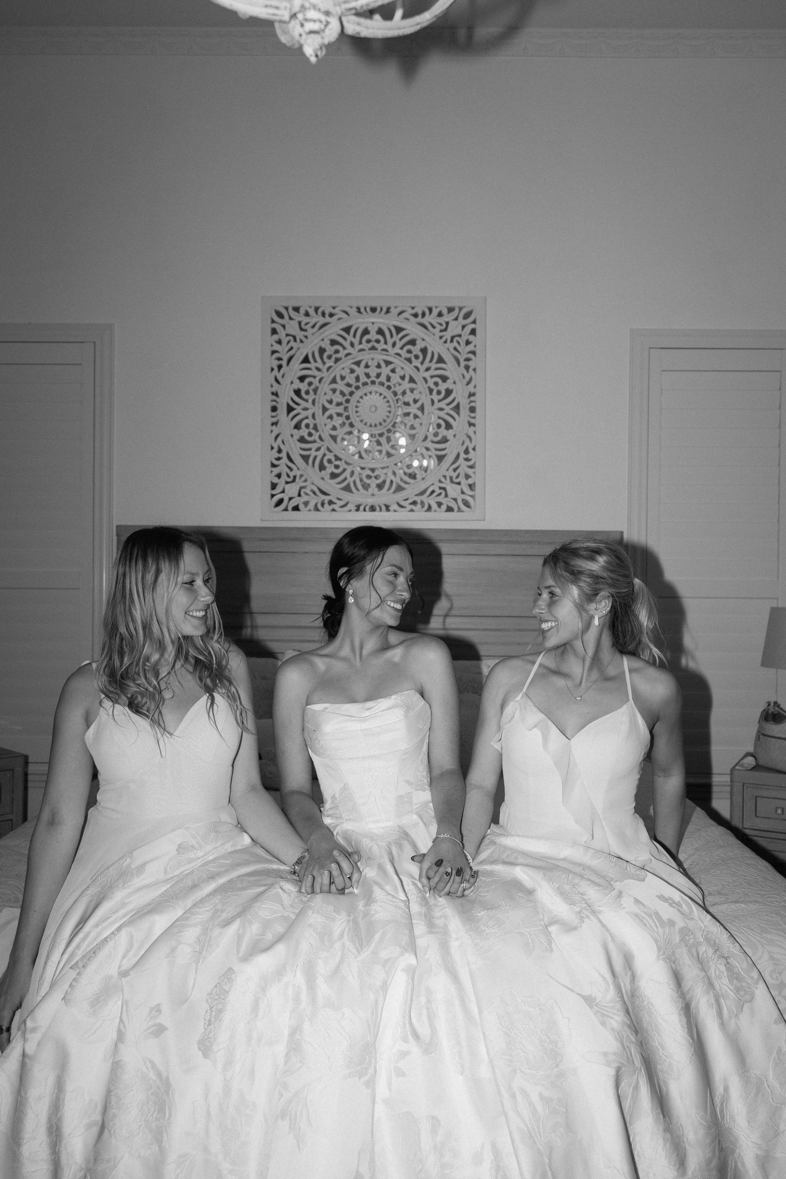 Three women in white wedding dresses sit together in a bright room with ornate wall decorations.