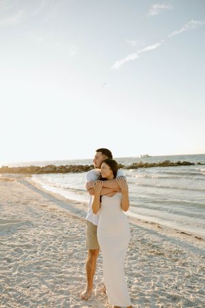 A couple embraces on a sandy beach during golden hour with ocean waves in the background.