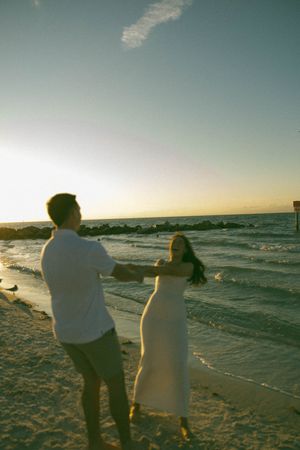 Two people stand on a beach at sunset as waves crash along the shoreline while golden light illuminates the scene.