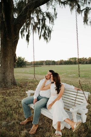 A couple sits together on a white wooden swing beneath a Spanish moss draped tree in a serene outdoor setting.