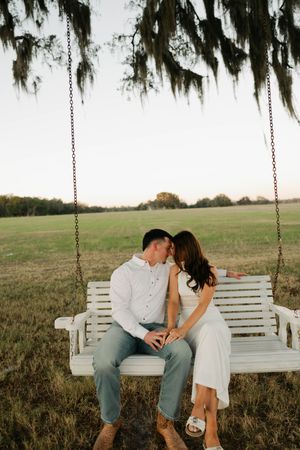 A romantic couple shares intimate moments on a white wooden porch swing in a rustic outdoor setting.