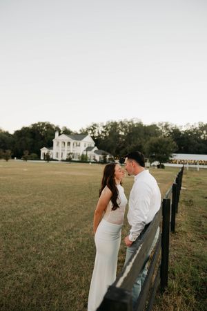 A romantic couple shares an intimate moment by a rustic fence with a white plantation-style house in the background.