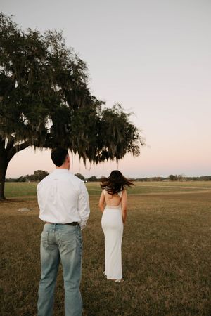 A couple stands in a field at sunset with Spanish moss hanging from a large oak tree.