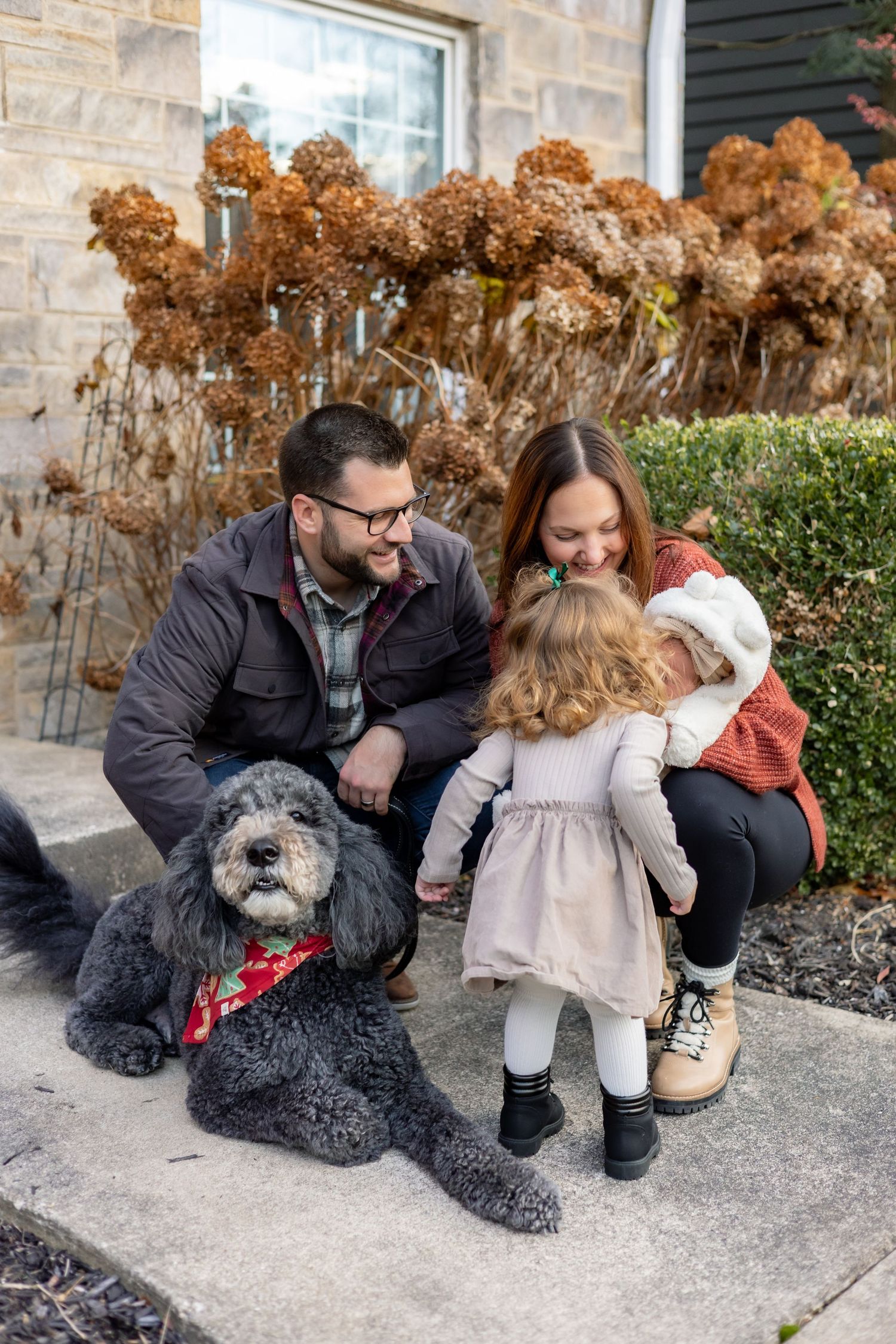 A couple sits outdoors with their fluffy gray dog against a stone wall with dried hydrangeas in autumn.