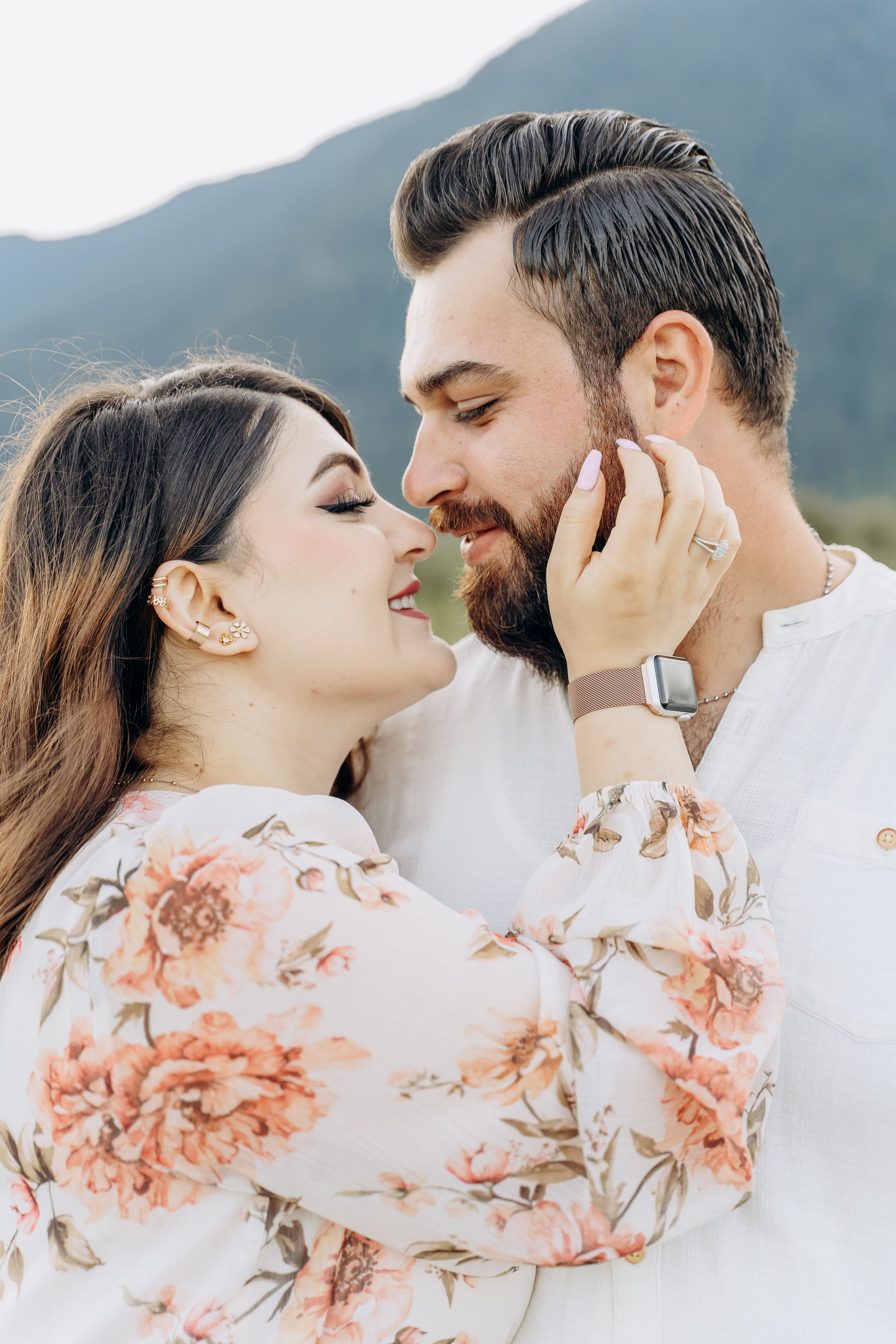 A romantic couple in white shares tender moments against a mountain backdrop with delicate pink floral details.