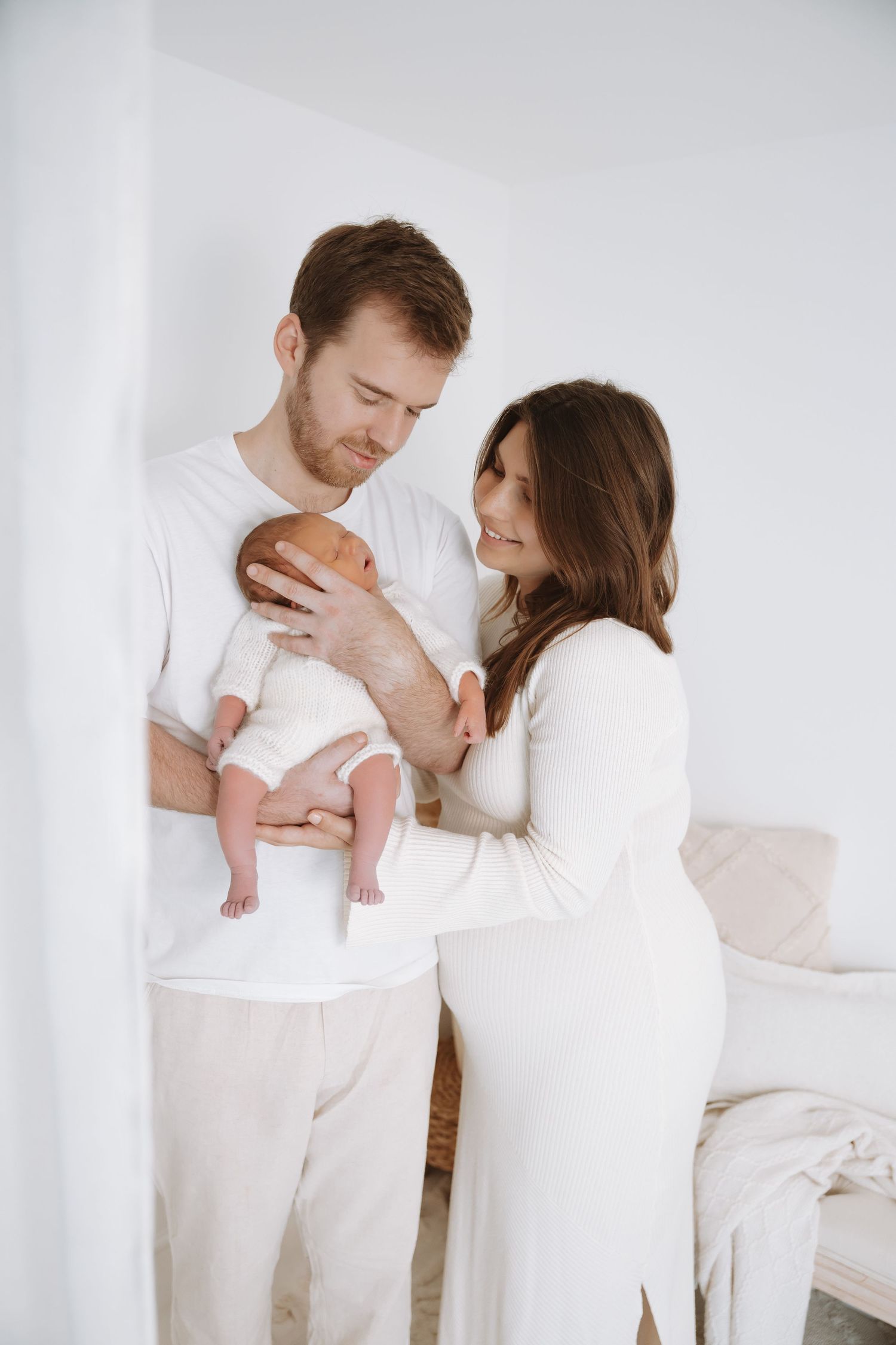 A couple in white clothing tenderly holds their newborn baby in a bright minimalist bedroom setting.