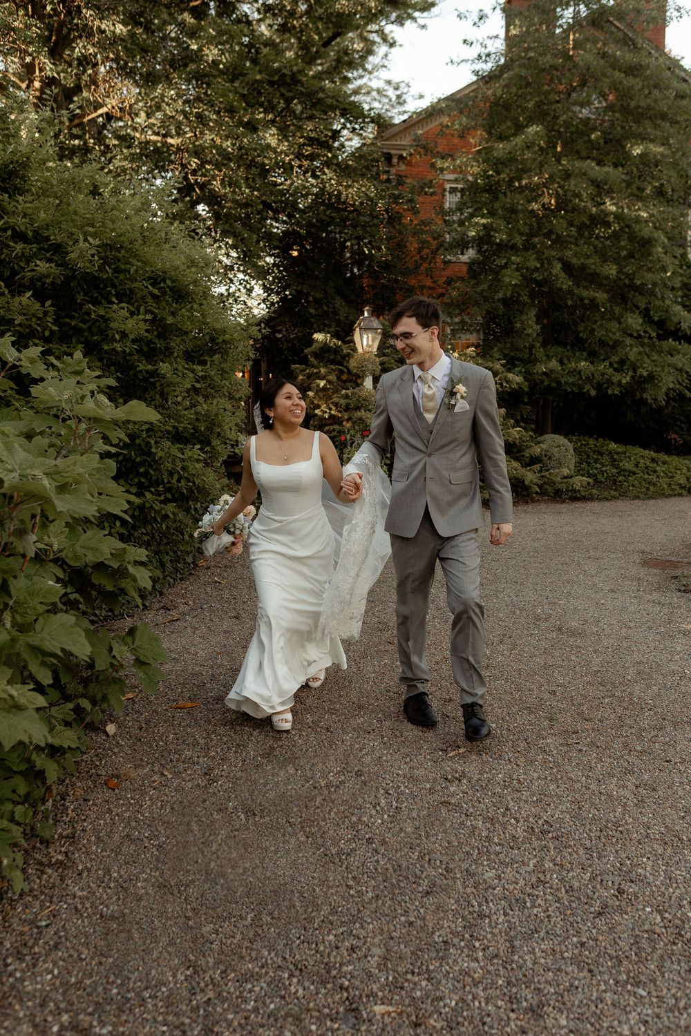 A couple in wedding attire runs playfully down a gravel path surrounded by lush green trees.