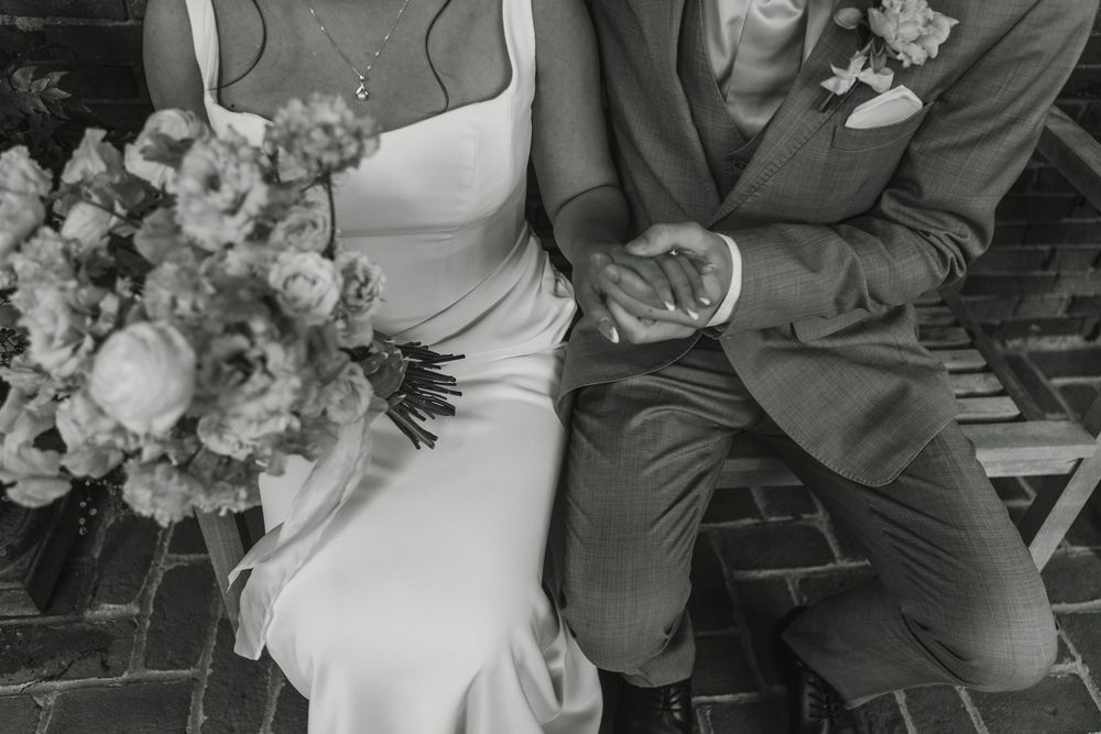 A wedding couple sits on wooden steps with their hands clasped together while surrounded by floral arrangements.