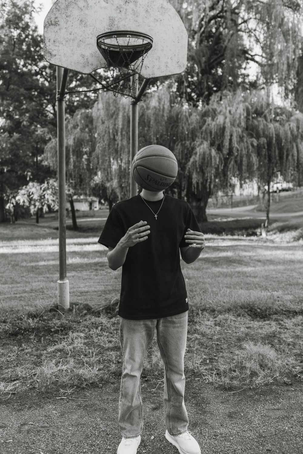 Black and white photo of someone balancing a basketball in front of their head at an outdoor court with willow trees.