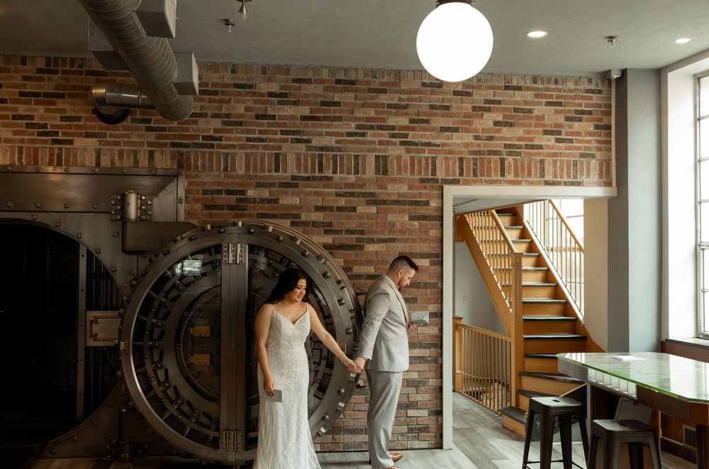 Wedding party poses in rustic brick-walled venue with natural lighting streaming through windows.