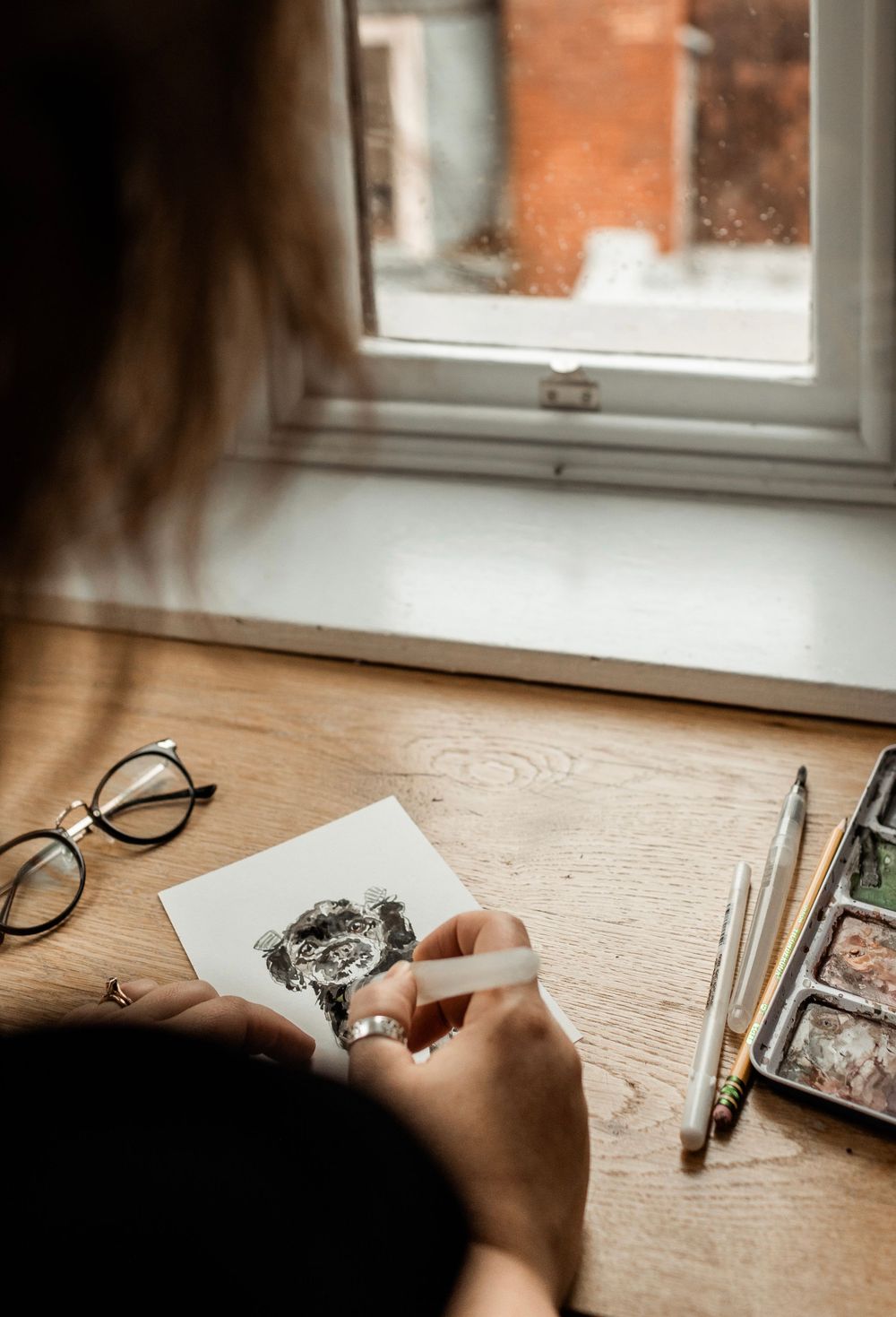 Artist sketches detailed portrait on wooden desk by window with natural lighting and art supplies.